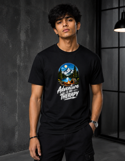 A man wearing a black short-sleeve t-shirt with a circular graphic of a mountain campsite and the text 'Adventure is Therapy' printed on the chest, standing in a dark industrial room.