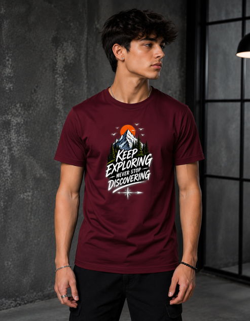 A man wearing a maroon crewneck t-shirt with a graphic print that reads 'KEEP EXPLORING never stop DISCOVERING' with a mountain and sun design, set against a dark gray textured studio background.