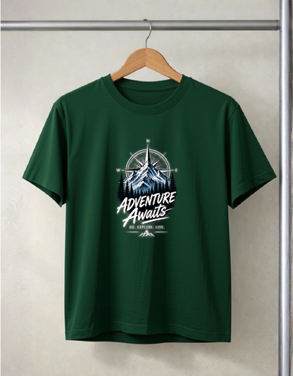 A Bottle green short-sleeve t-shirt hanging on a wooden hanger against a light gray wall. The shirt features a graphic print on the chest with a mountain, compass rose, and the text 'ADVENTURE Awaits' in white and light blue.