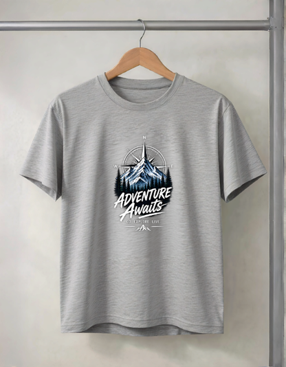 A heather grey Melange short-sleeve t-shirt hanging on a wooden hanger from a metal clothing rack. The shirt features a graphic print in the center with a mountain illustration and the text 'ADVENTURE AWAITS' in white script.