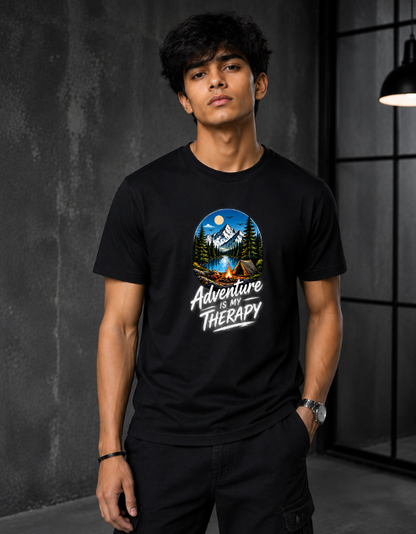 A man wearing a black short-sleeve t-shirt with a circular graphic of a mountain campsite and the text 'Adventure is Therapy' printed on the chest, standing in a dark industrial room.