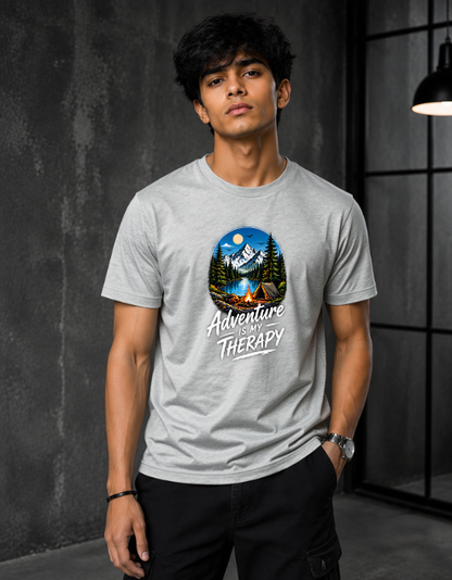 A man wearing a gray Melange crewneck t-shirt with a circular camping graphic that reads 'Adventure is Therapy'. The background is a dark gray textured wall with a black metal grid window and a hanging lamp.
