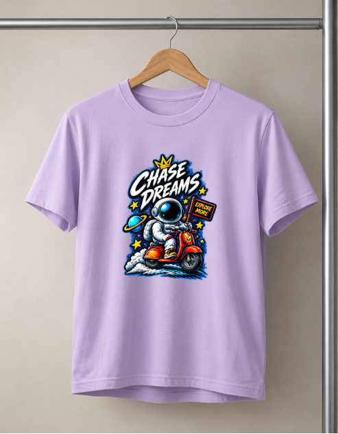 A Lavender color crewneck t-shirt hanging on a wooden hanger against a neutral beige wall. The shirt features a colorful graphic of an astronaut riding an orange scooter with the text 'Chase Dreams' and stars.