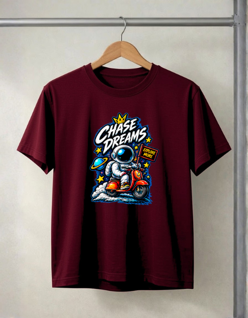 A maroon short-sleeve t-shirt hanging on a wooden hanger against a light gray wall. The shirt features a colorful retro graphic of an astronaut riding an orange scooter with the text 'Chase Dreams' in white and blue.