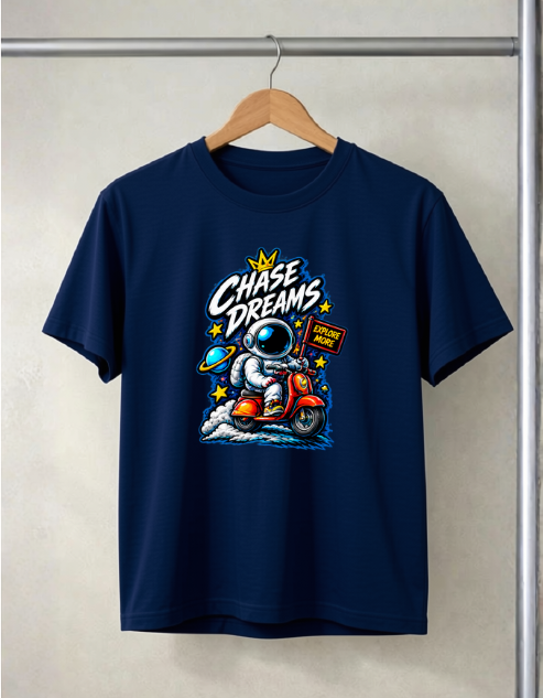 A navy blue short-sleeve t-shirt hanging on a wooden hanger against a light beige wall. The shirt features a colorful graphic print of an astronaut riding a red scooter with the text 'Chase Dreams' and a small sign that reads 'I Want To Fly'.