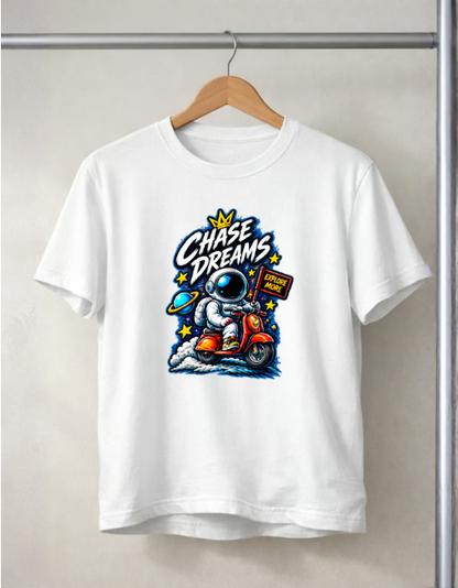 A white short-sleeve crewneck t-shirt hanging on a wooden hanger against a light beige wall. The shirt features a colorful graphic print of an astronaut riding a red scooter with the text 'Chase Dreams' and stars.