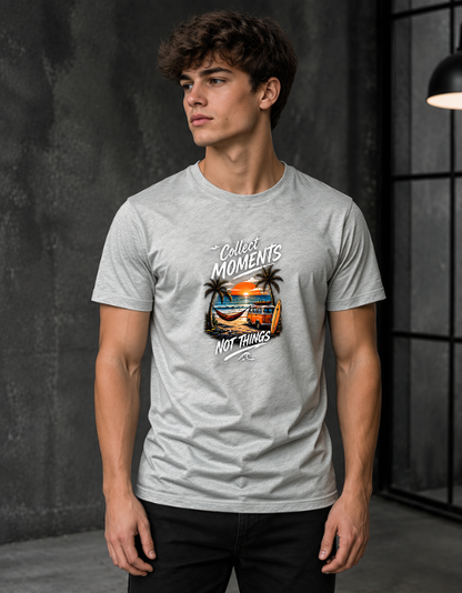 A man wearing a light grey Melange heather t-shirt with a graphic print that reads 'Collect MOMENTS NOT THINGS' around a beach sunset illustration, standing in a dark gray studio setting.