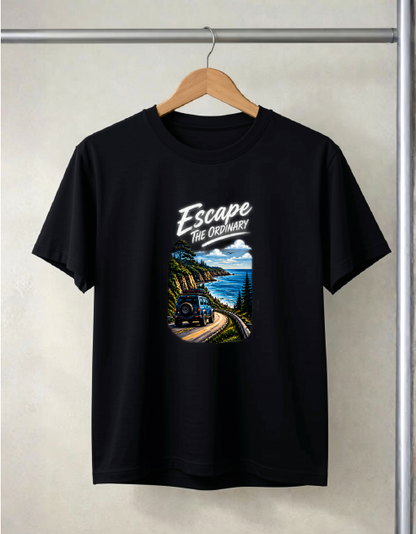 A Black color short-sleeve t-shirt hanging on a wooden hanger against a light gray wall. The shirt features a graphic print on the front with the text 'Escape The Ordinary' above an illustration of a train traveling along a scenic coastal mountain road.