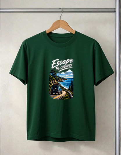 A Bottle green short-sleeve t-shirt hanging on a wooden hanger against a light gray wall. The shirt features a graphic print on the front with the text 'Escape The Ordinary' above an illustration of a train traveling along a scenic coastal mountain road.