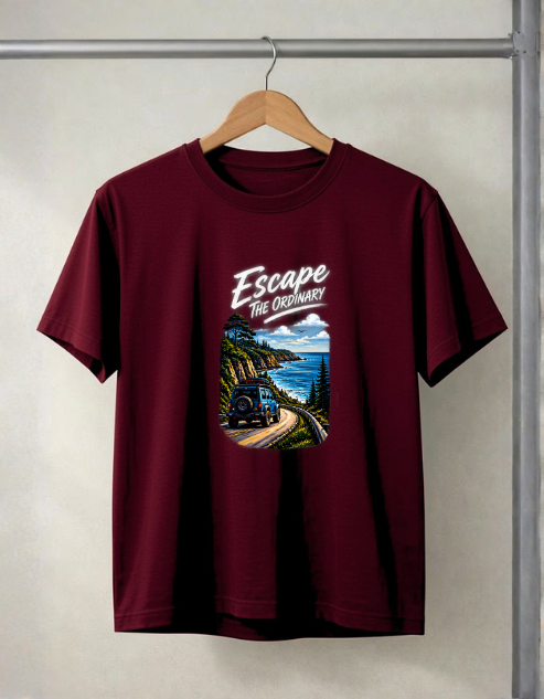 A maroon short-sleeve t-shirt hanging on a wooden hanger from a metal clothing rack. The shirt features a graphic print on the front with the text 'Escape The Ordinary' above an illustration of a vintage camper truck traveling along a winding coastal road.