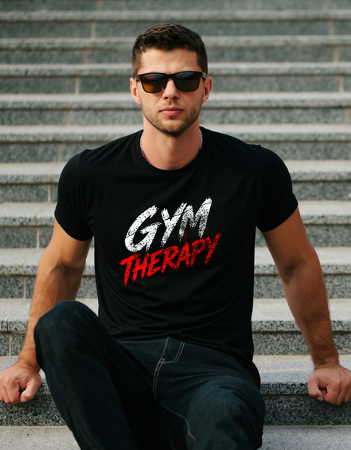 A man wearing a black t-shirt with 'GYM THERAPY' printed on the chest, sitting on outdoor stone steps. The text on the shirt features 'GYM' in a textured white brushstroke font and 'THERAPY' in a distressed red brushstroke font.