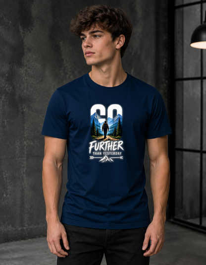 A man wearing a navy blue graphic t-shirt with a white and multicolor outdoor print that reads 'GO FURTHER THAN YESTERDAY'. The photo is taken in a dimly lit, modern industrial loft setting with a dark textured wall.