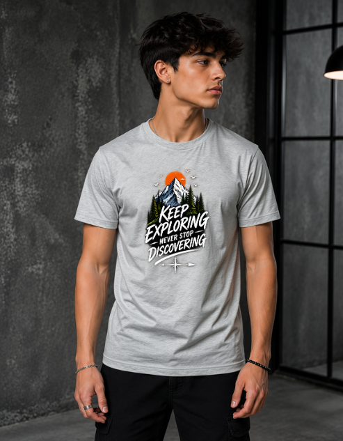 A man wearing a light gray crewneck t-shirt with a graphic print that reads 'KEEP EXPLORING WHERE YOU'RE DISCOVERING' around a mountain and sunset illustration. He is standing in a dark, modern industrial room with a textured gray wall.