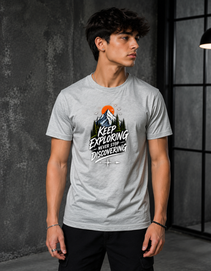 A man wearing a light gray crewneck t-shirt with a graphic print that reads 'KEEP EXPLORING WHERE YOU'RE DISCOVERING' around a mountain and sunset illustration. He is standing in a dark, modern industrial room with a textured gray wall.