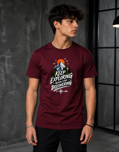 A man wearing a maroon crewneck t-shirt with a graphic print that reads 'KEEP EXPLORING never stop DISCOVERING' with a mountain and sun design, set against a dark gray textured studio background.
