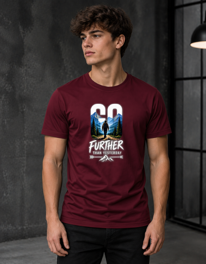 A man wearing a maroon short-sleeve graphic t-shirt with a 'GO FURTHER THAN YESTERDAY' outdoor hiker design, standing in a dark industrial loft setting.