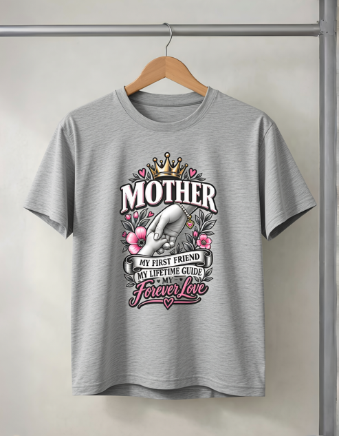 A light gray heather crewneck t-shirt hanging on a wooden hanger against a neutral wall. The shirt features a graphic print on the front with the word 'MOTHER' in bold white letters under a gold crown, surrounded by pink flowers, a white hand, and decorative banners reading 'MY FIRST FRIEND', 'MY LIFETIME GUIDE', and 'Forever love'.