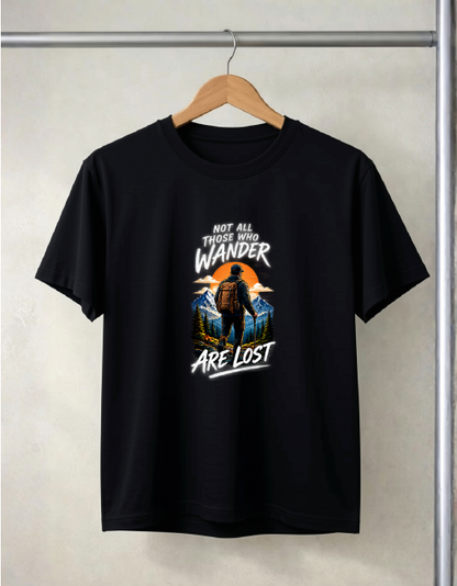 A black short-sleeve t-shirt hanging on a wooden hanger against a light beige wall. The shirt features a graphic print on the front with a silhouette of a person hiking against an orange sunset, with the text 'NOT ALL THOSE WHO WANDER ARE LOST' in white.