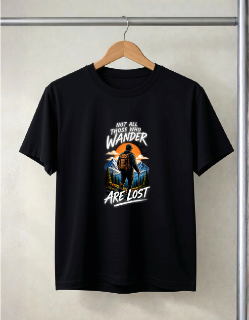 A black short-sleeve t-shirt hanging on a wooden hanger against a light beige wall. The shirt features a graphic print on the front with a silhouette of a person hiking against an orange sunset, with the text 'NOT ALL THOSE WHO WANDER ARE LOST' in white.
