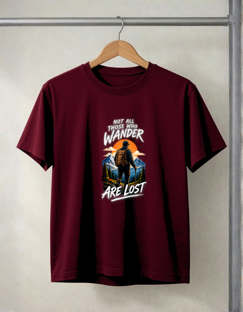A maroon short-sleeve t-shirt hanging on a wooden hanger against a light gray wall. The shirt features a graphic print on the front with the text 'NOT ALL THOSE WHO WANDER ARE LOST' surrounding an illustration of a person hiking against a sunset and mountain backdrop.
