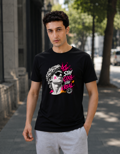 A man wearing a black graphic t-shirt with a vibrant pink, yellow, and white design featuring a woman's portrait and the text 'STAY COOL SAY REAL' in a graffiti-style font, standing on a city sidewalk.