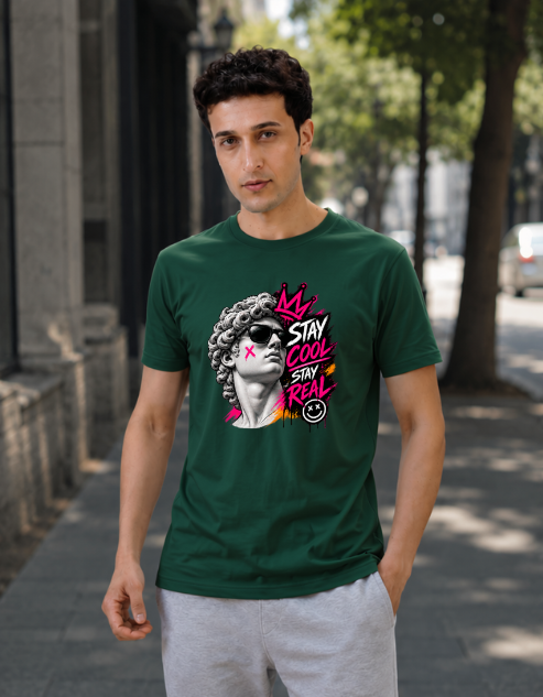 A man wearing a navy bottle green graphic t-shirt with a stylized portrait and the text 'STAY COOL SAY REAL' in pink, white, and yellow, standing on a city sidewalk.